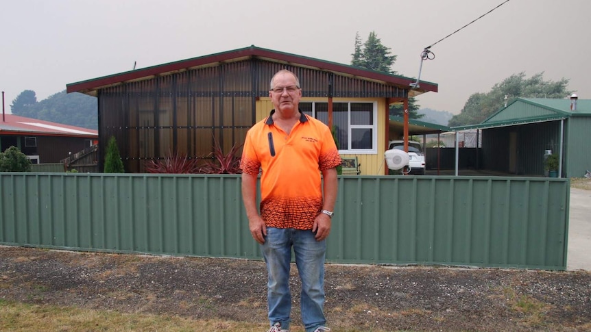 A man stands in front of a home in Zeehan, in Tasmania. It's smoky in the background.
