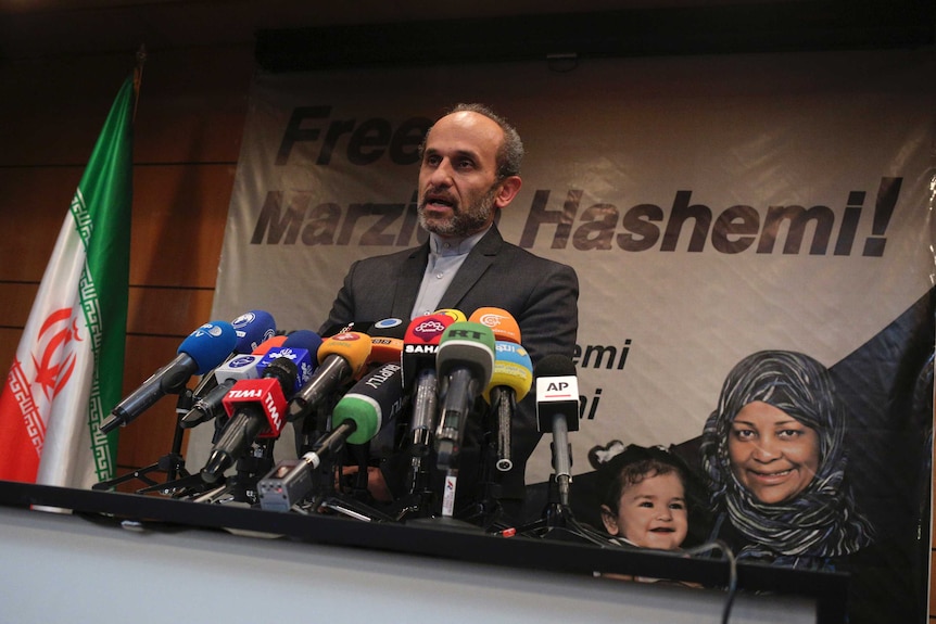 A man gives a press briefing in front of a poster saying 'Free Marzieh Hashemi'.