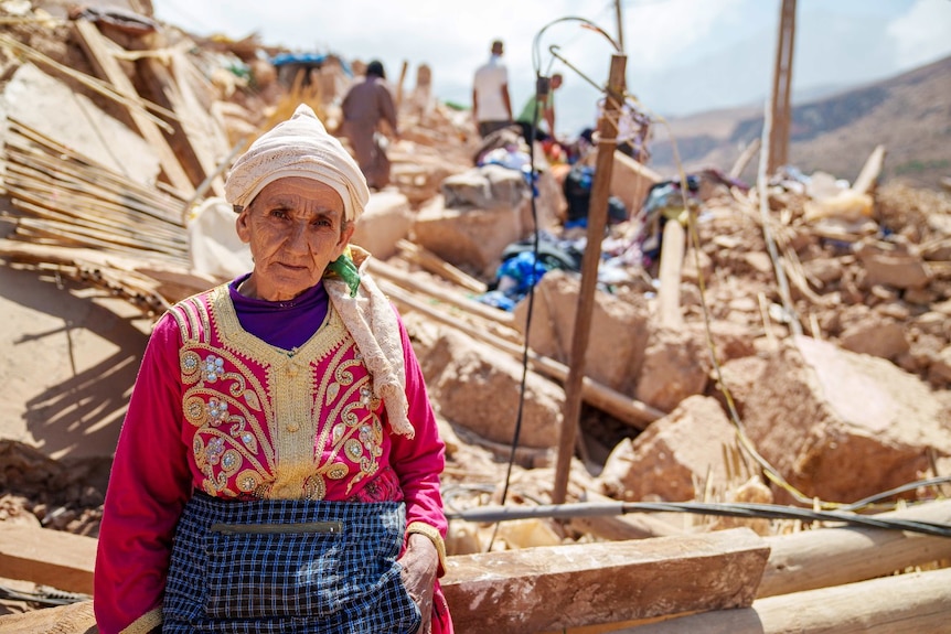 A woman looks at the camera, with a destroyed building visible in the background.