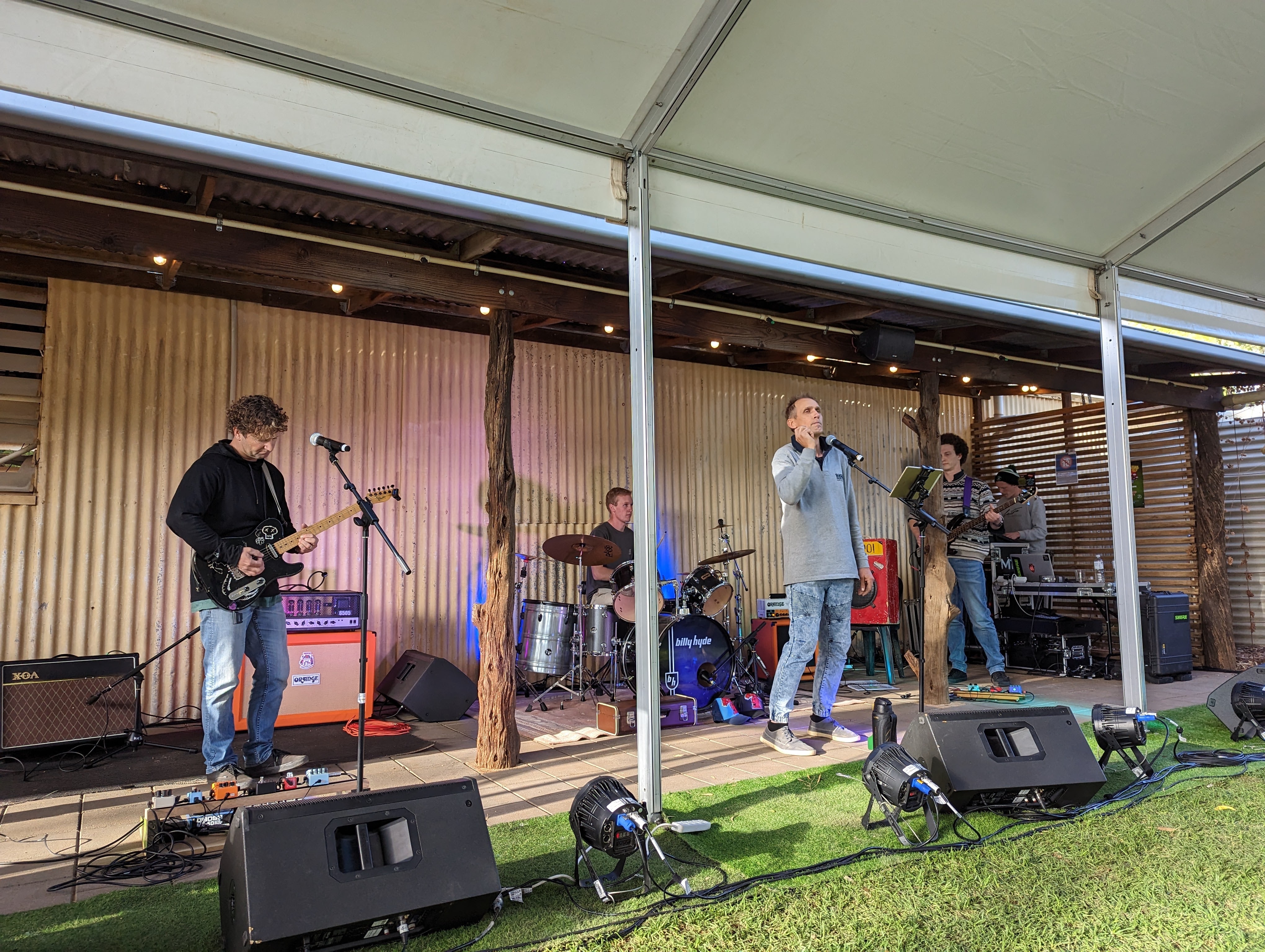 Four men standing under a shade playing instruments and singing