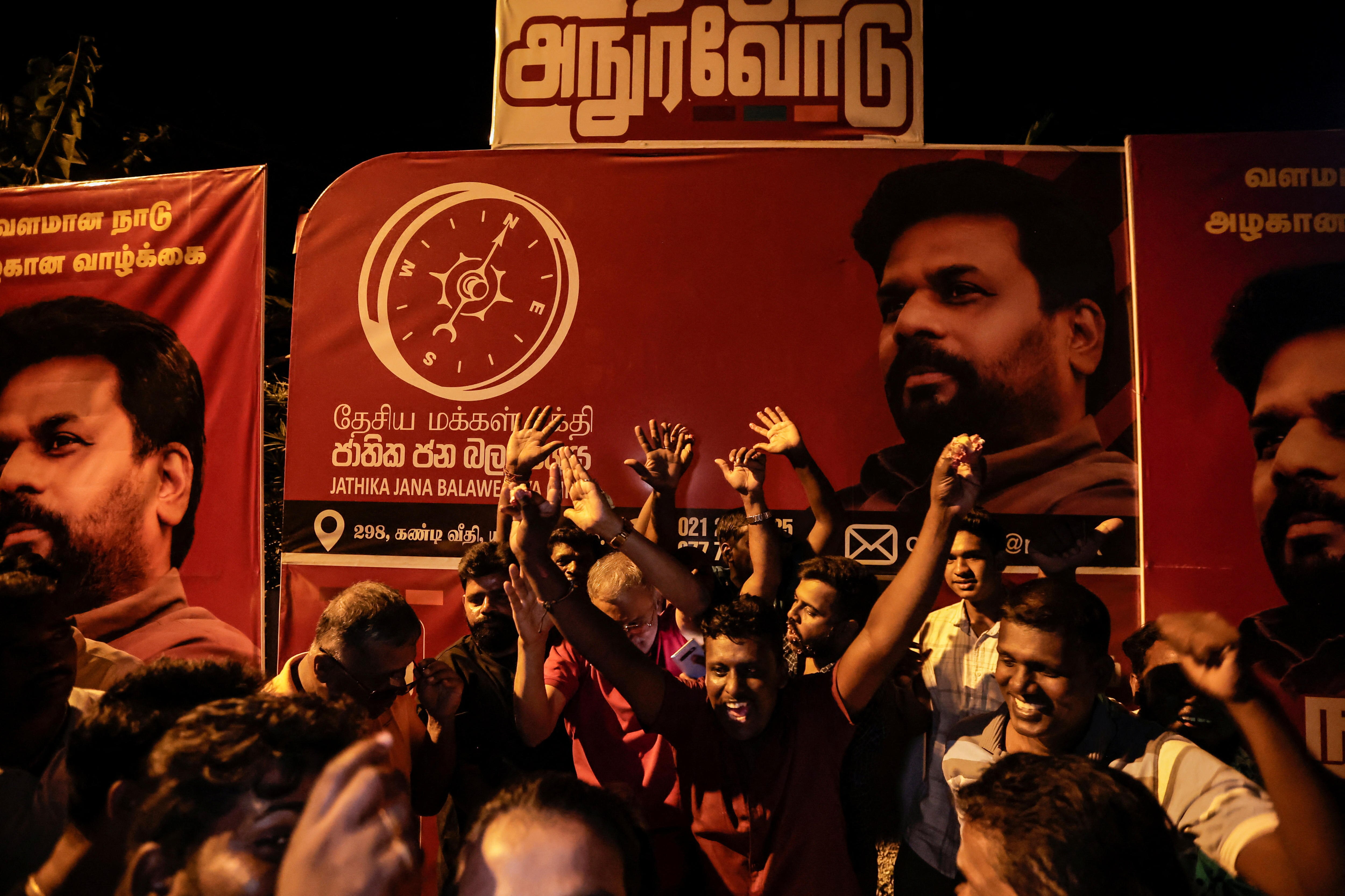 A group of people stand in front of a picture of Anura Dissanayake cheering and smiling