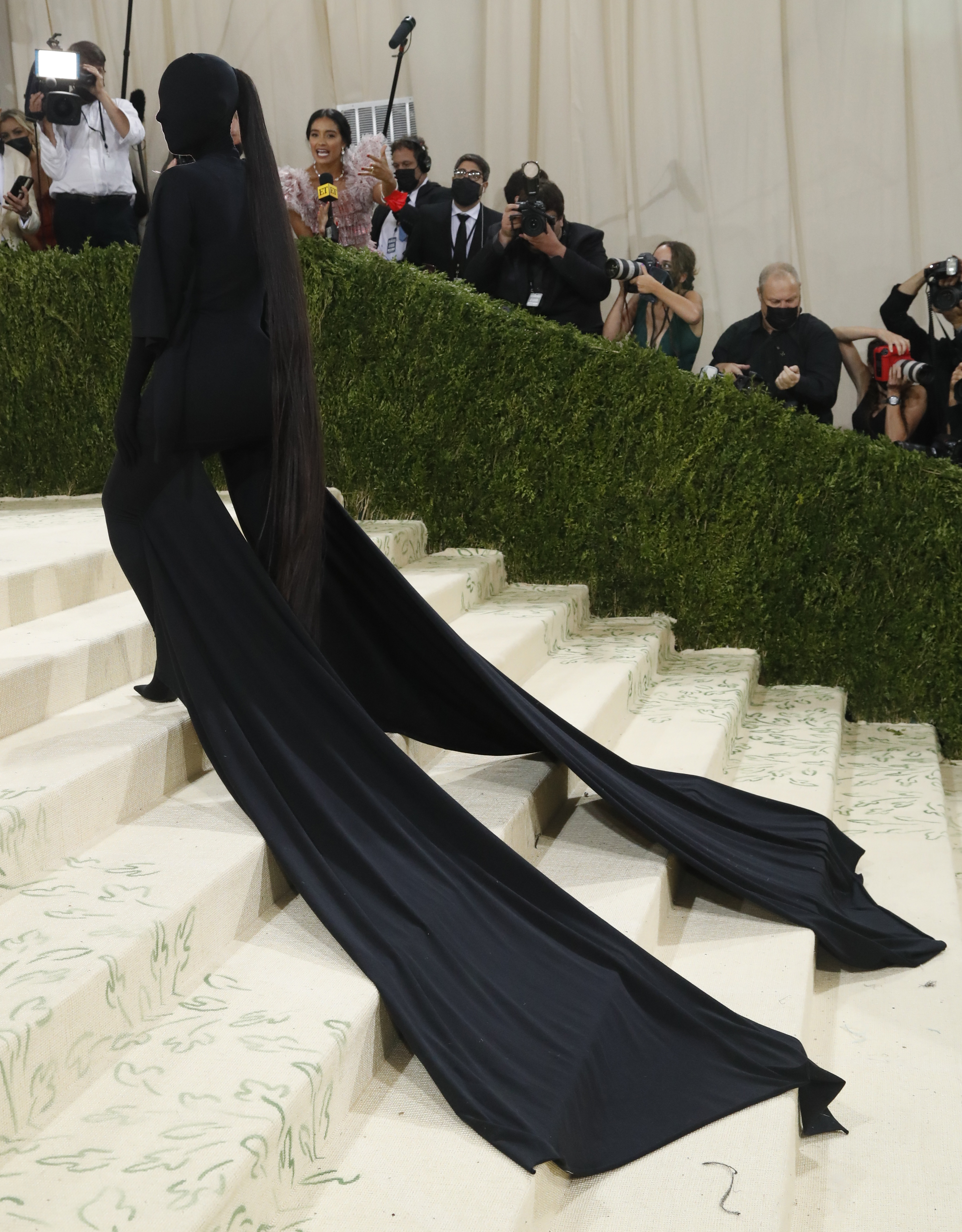 Kim Kardashian wearing a full black fitted suit, covering her face. a long black ponytail and caped sleeves trail behind her. 