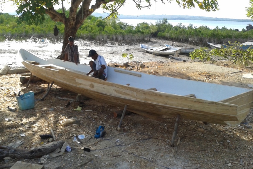 a man paints a wooden boat under a shady tree with a decayed  boat in the background