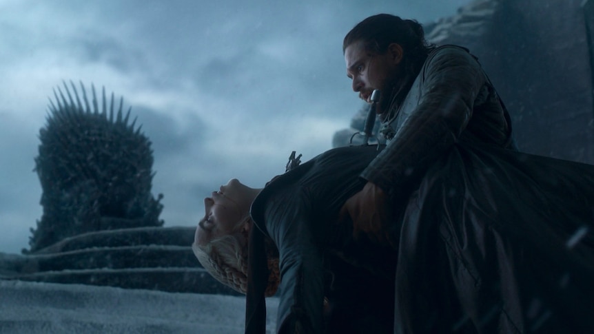 Daenerys lies limp in Jon's arms with the Iron Throne in the background.