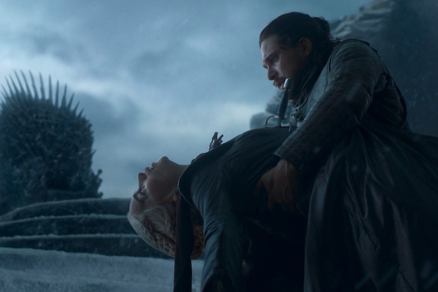 Daenerys lies limp in Jon's arms with the Iron Throne in the background.