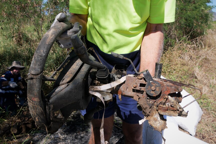 A person wearing a high visibility work shirt and gloves holds a rusted steering wheel in bushland.