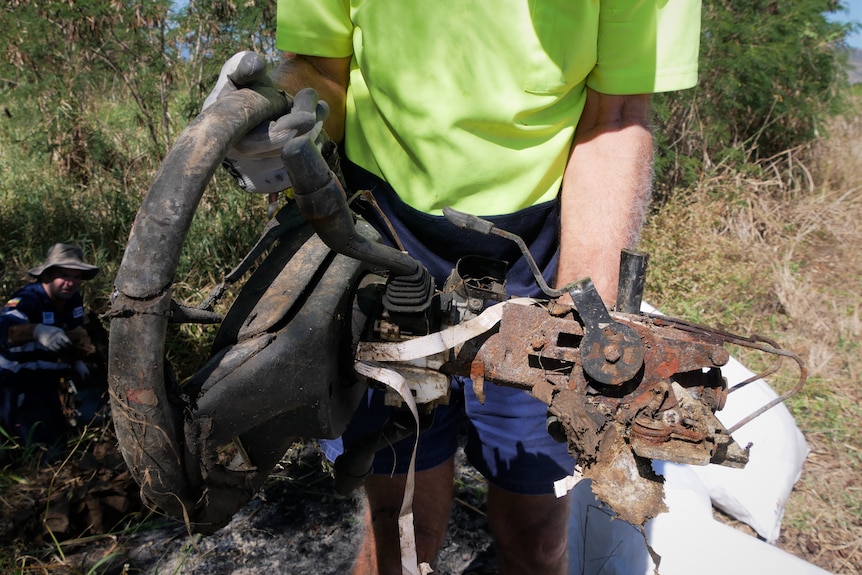 A person wearing a high visibility work shirt and gloves holds a rusted steering wheel in bushland.