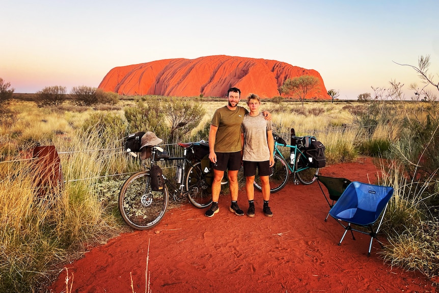 A father and son stand in front of Uluru with their bicycles.
