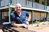 A man in a blue polo and bald head smiles while leaning against a wooden bench with a house behind