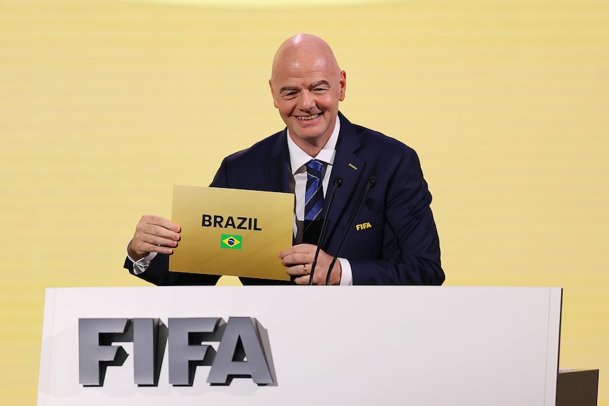 A bald man wearing a suit holds a gold-coloured card saying "Brazil" at a podium
