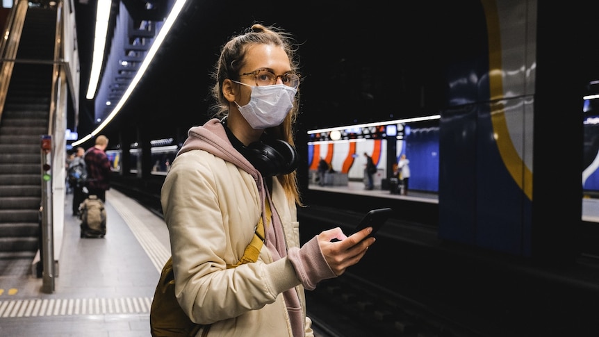 A young woman wearing a mask checks her phone while waiting for a train