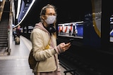 A young woman wearing a mask checks her phone while waiting for a train