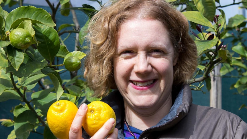 A woman stand in front of a lemon tree, holding lemons