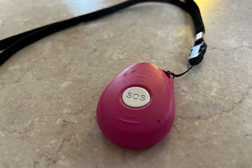 A pink medical alert pedant with a silver SOS button and a black strap sitting on a white table top.