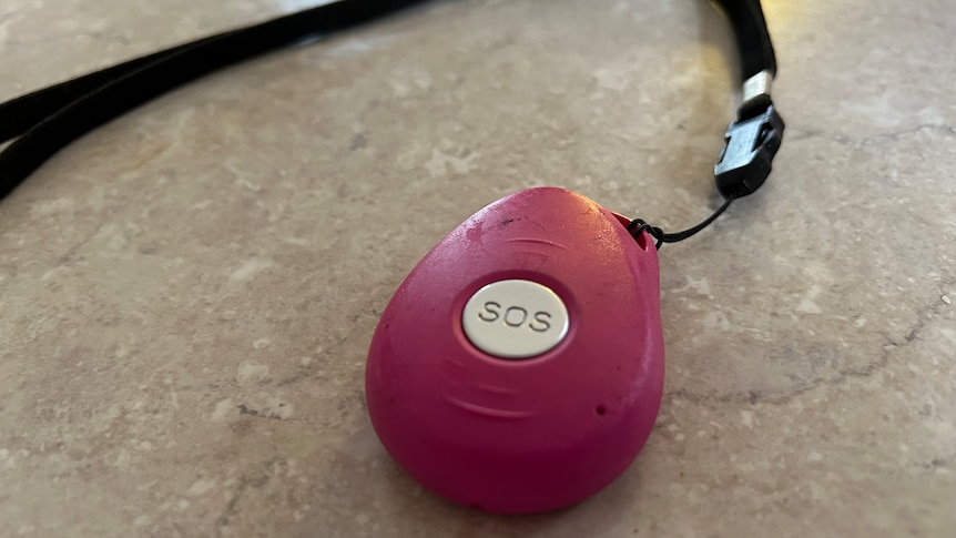 A pink medical alert pendant with a silver SOS button and a black strap sitting on a white table top.