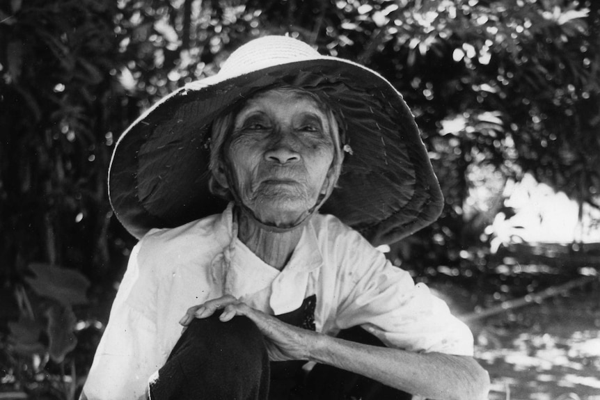 Elderly Chinese lady wearing a wide-brimmed hat crouching in her garden holding a machete looking at the camera