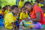 A group of children surround a woman writing in a notebook