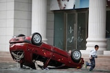 A boy runs away from a red car overturned on the street.