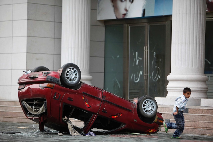 A boy runs away from a red car overturned on the street.