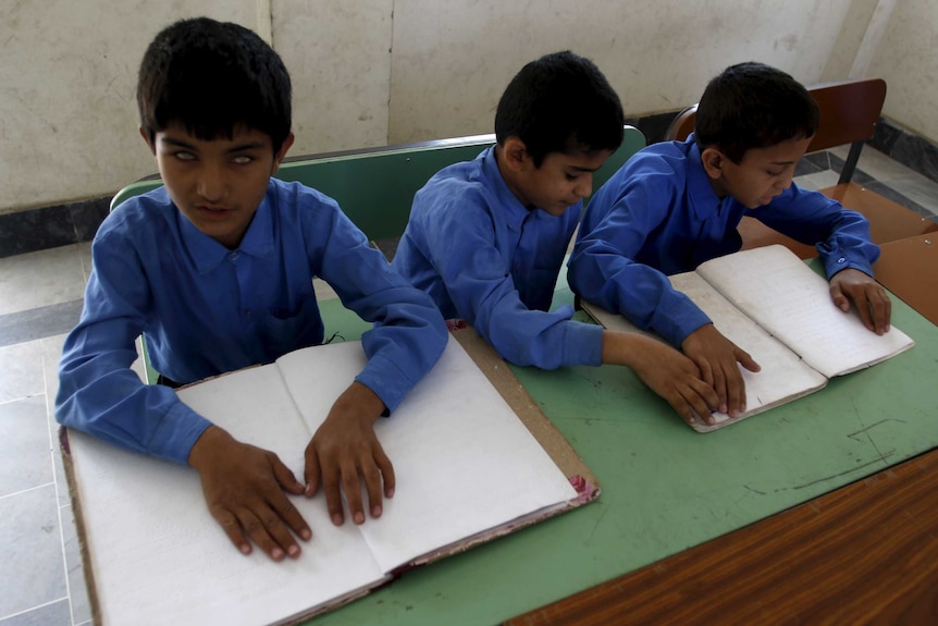 Three blind children in blue shirts sit at green desk and run their hands over open exercise books.