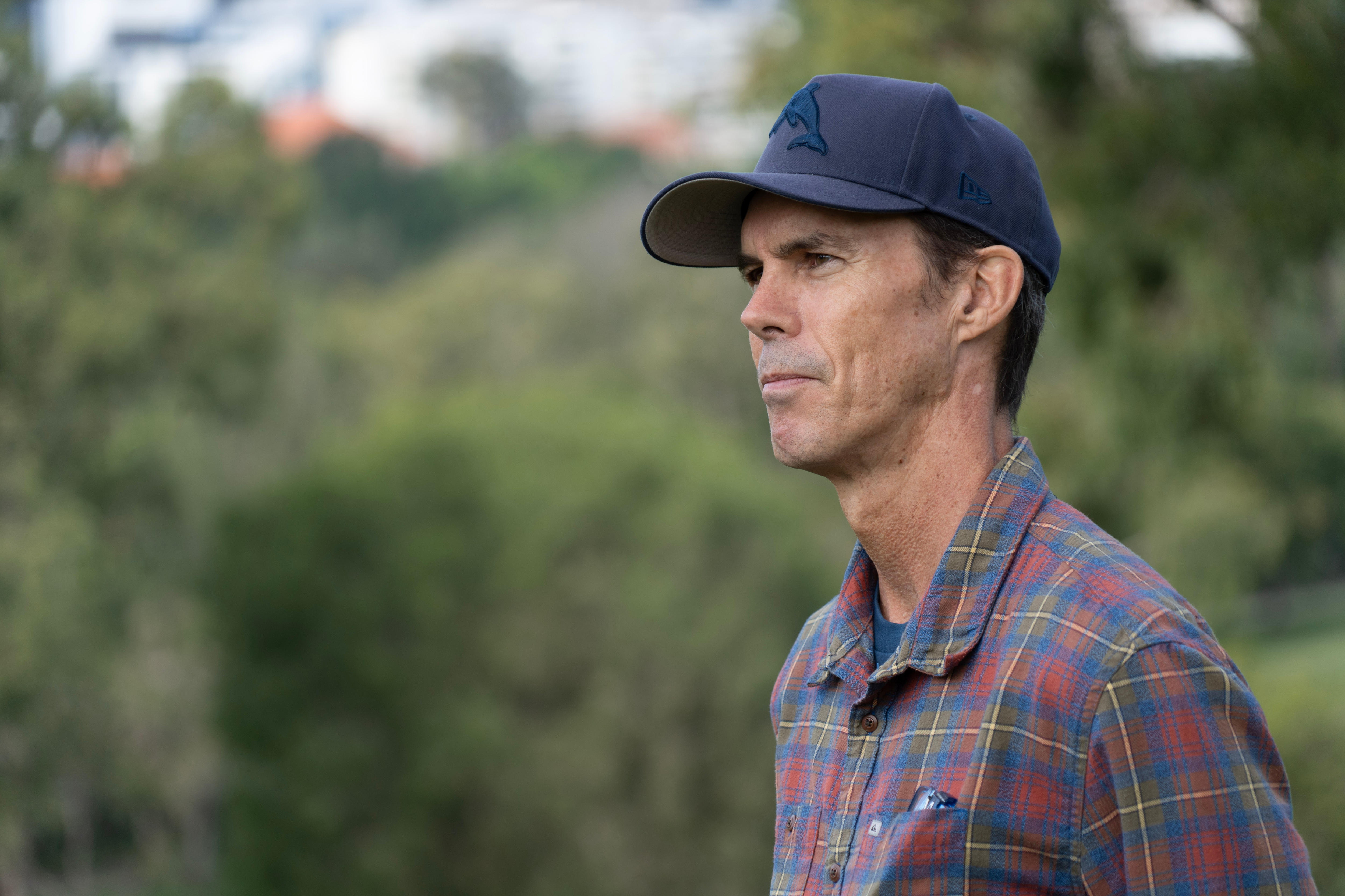 A man in a cap and chequered shirt looking to right of frame with tree in background.