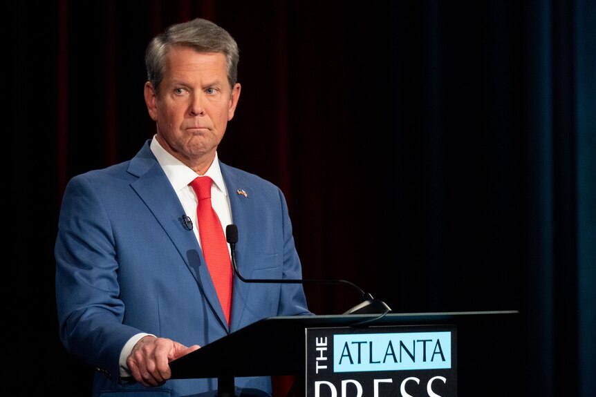 Governor Brian Kemp on stage at the Atlanta Press Club wearing a blue suit and red tie