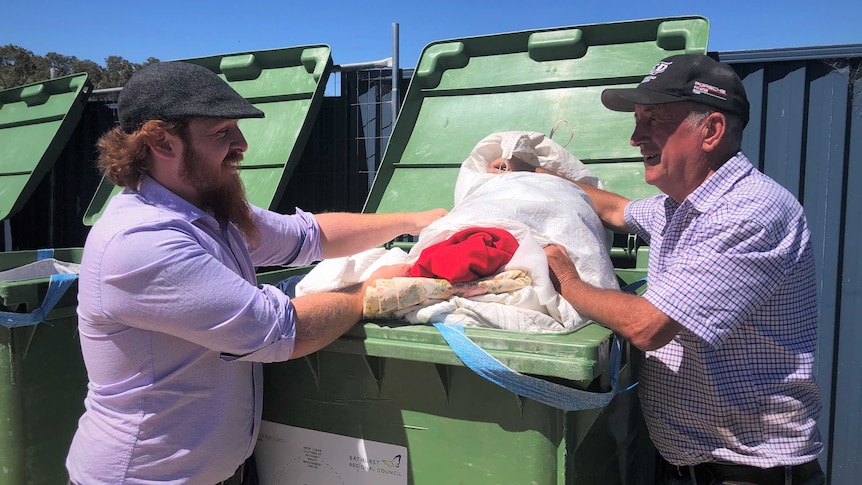 Two men standing sideways in front of a waist-high green bin full of clothes.