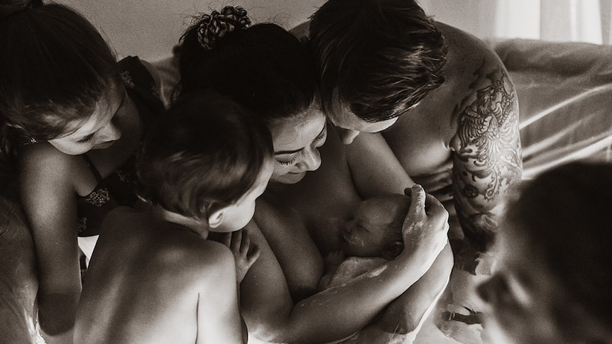 Children and a man gather closely around a mother who hugs her newborn while sitting inside in a shallow birth pool.