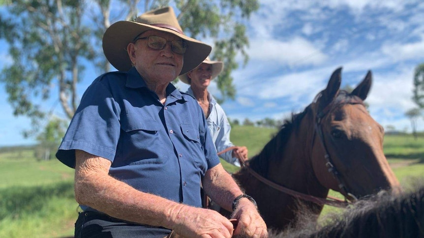 An elderly man in a blue shirt and hat rides a horse.