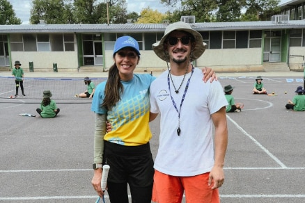 Two people, a woman and man, stand arm in arm on the tennis court wearing hats and holding racquets, smiling.