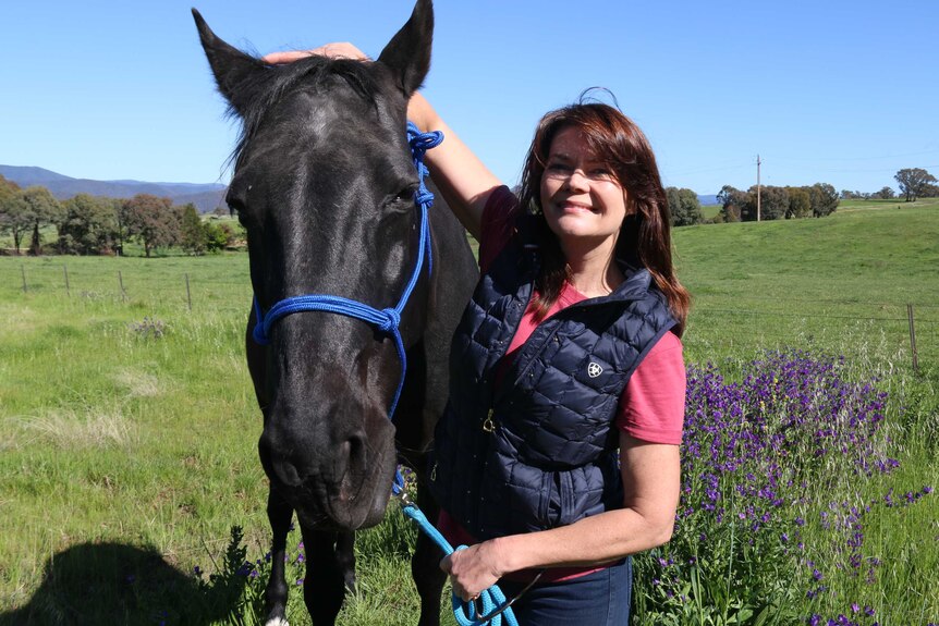 A smiling woman patting a shiny black horse in a green field, with clumps of purple Patterson's curse in the background.