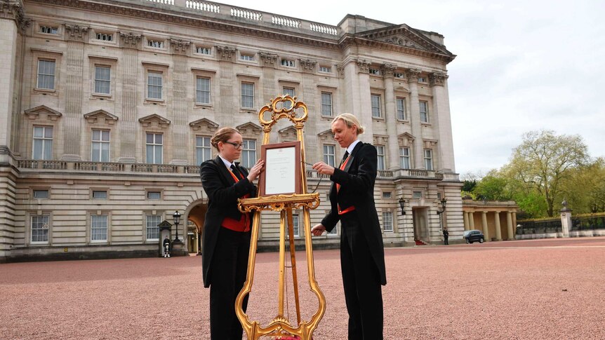 Two stewards add an announcement to an easel in front of Buckingham Palace