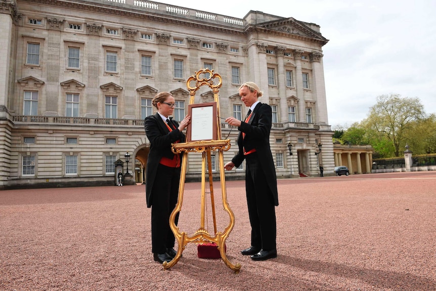 Two stewards add an announcement to an easel in front of Buckingham Palace