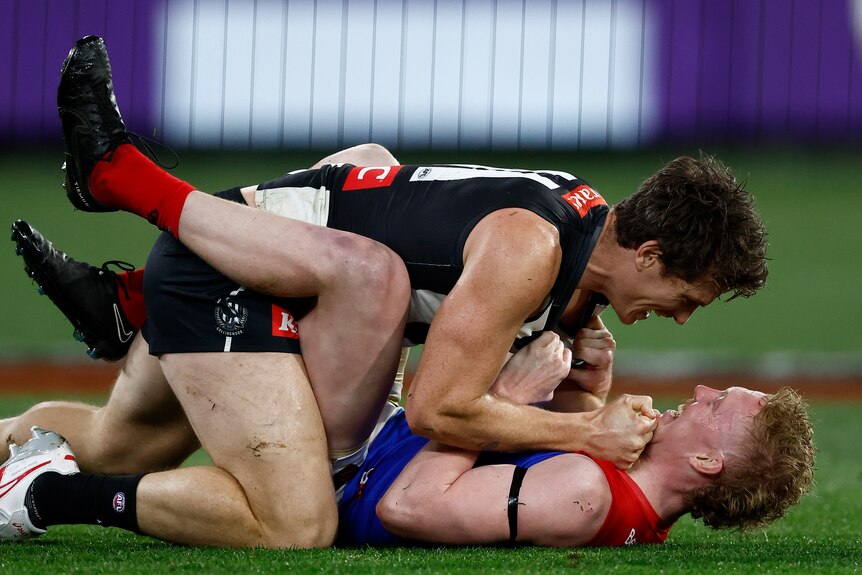 Two AFL players wrestle on the ground, one in black and white on top of one in red and blue