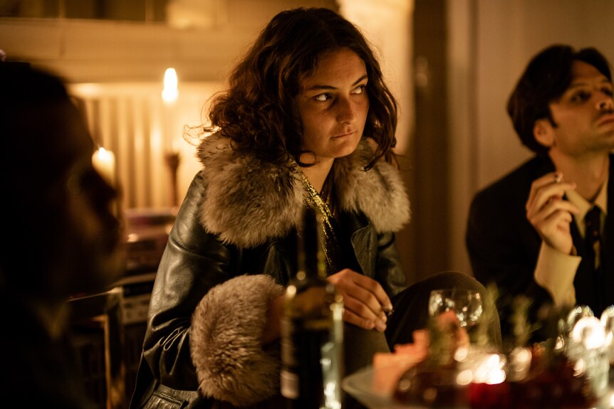 A woman in her mid 20s with dark hair and leather and fur jacket sits at table, other people around her