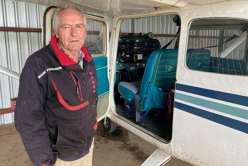 An older man stands in a shed, next to a small plane with its door open.