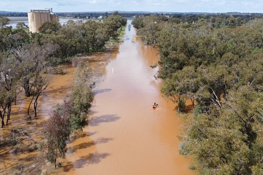 An aerial view of a road covered with brown floodwater, and a kayak with two teenagers paddling.