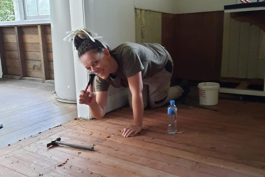 A woman kneeling down and holding tools to use on the floor of her empty home.