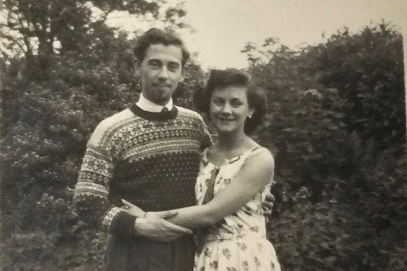 A black and white photograph shows a young man and woman standing together with arms around each other in a garden
