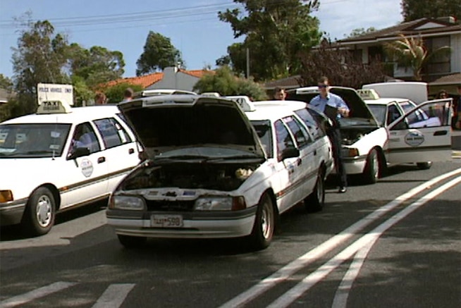 A policeman and another man stand at the rear of a taxi with its bonnet raised, with other taxis surrounding them.