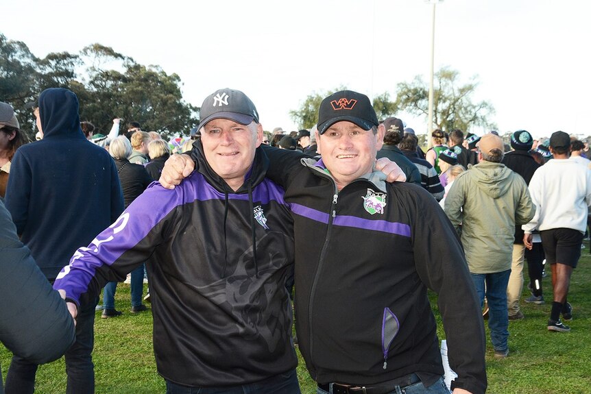 Two men in black and purple coats embrace on a crowded football field. The one on the left is crying.