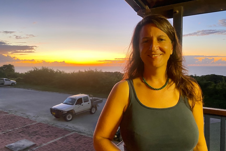 A woman in a green top stands in front of a sunset. 