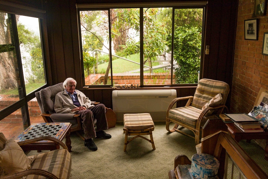 An older man sits in his house in Grafton, with the river behind him.