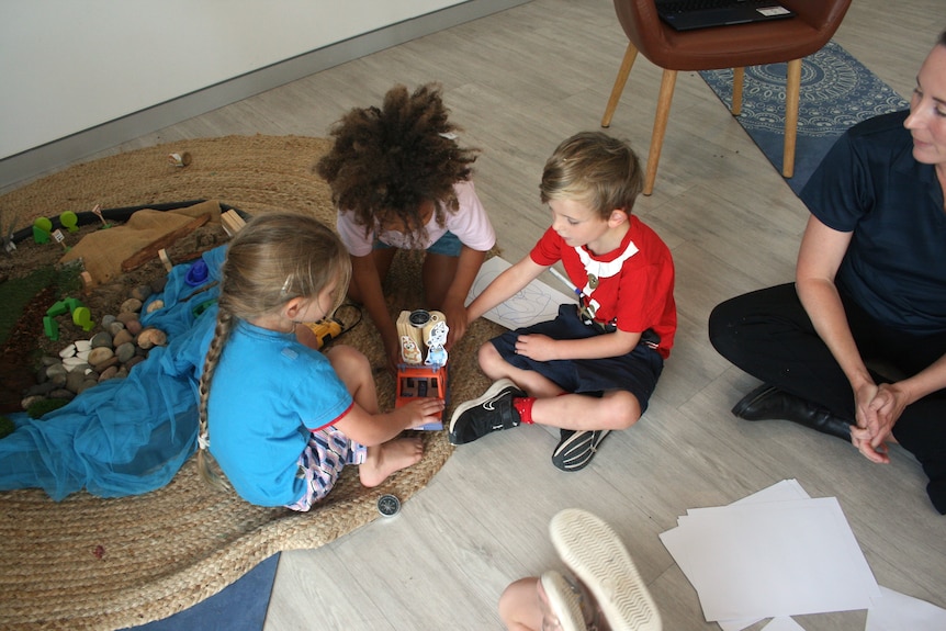 Three children play with a toy together in a circle while an adult watches on.