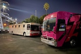 Front of large pink bus in at night under lights with a smaller, white and pink bus behind it