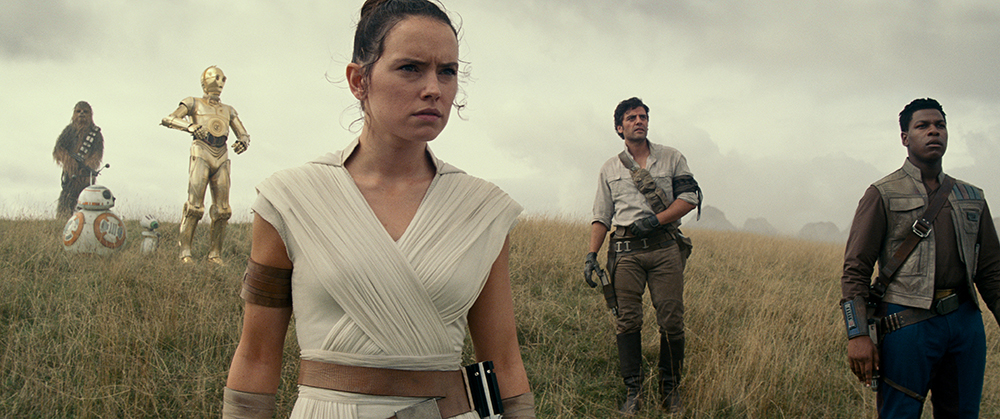 Chewbacca, two small droids, C-3PO, Daisy Ridley, Oscar Isaac and John Boyega stand in dry grassland with serious expressions.