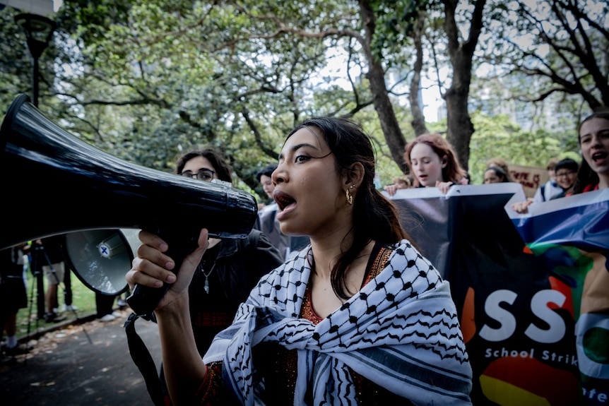 A girl with dark hair, wearing a Keffiyeh around her shoulders, shouts into a megaphone, students march behind.