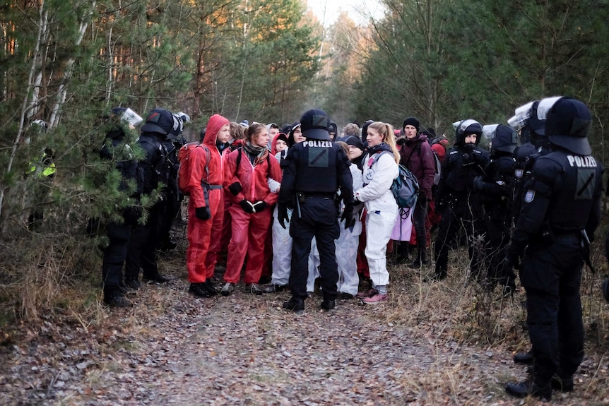 Protesters in white and red jumpsuits are blocked by police in black riot gear.