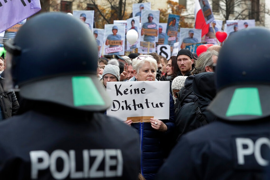 From behind a line of riot police, you view a crowd with a woman holding a sign in German that reads 'no dicatorship'.