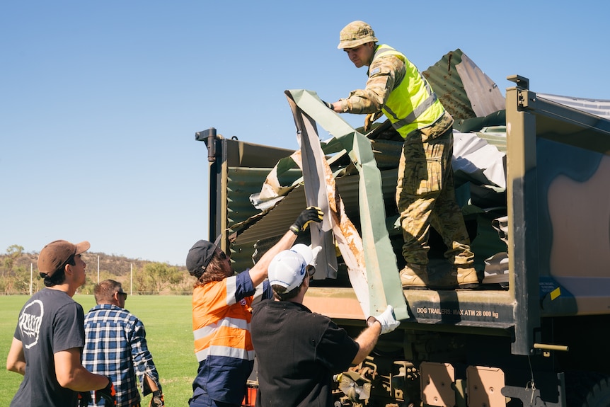 An Army reservist in camouflage fatigues stands on a truck lifting debris into the back while helping four men on the ground. 